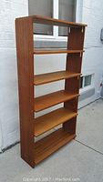 Full side view of teak bookcase standing outside against a white brick wall, showing the five shelves and open design.