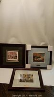 Three framed prints displayed, showing varied wood frames and mats. Two upright and one flat. Corner protectors on two frames.