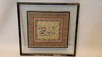 Framed square handkerchief with oriental embroidery, hung on a white wall.