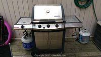 Full view of the stainless steel propane BBQ grill with side burner, control knobs, front cabinet, and propane tanks on each side.