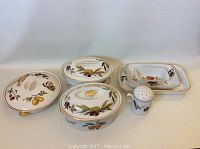 Top view of covered oval and round baking dishes, rectangular platters and pinch shaker, all with fruit designs and gold accents.