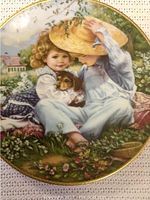 Close-up of 'Reaching Together' plate depicting two children and a dog, one holding a straw hat, with detailed greenery and a small house in background. Plate rimmed with gold.