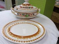 Photo of ceramic soup tureen with handles and lid, decorated with vegetables and gold scallop border, alongside matching oval platter.