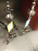 Pair of tarnished metal andirons placed on a concrete floor, showing full view of both andirons with tags attached.