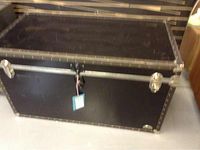 Front view of a large black trunk with metal reinforcements and latches, placed on the floor.