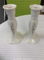 Pair of cream-colored Belleek candle sticks with floral design, shown on white cloth background with green pegboard behind.