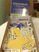 Photo showing a full blue and yellow Statesman Deluxe Album with gold lettering, loose stamps and small yellow envelopes inside a box.