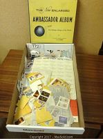 Photo of an open white box containing loose postage stamps and a partially visible stamp album, titled The New Enlarged Ambassador Album