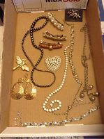 Top view of assorted jewelry pieces laid out in a box, showing various necklaces, brooches, bracelets, and earrings including a heart-shaped rhinestone brooch and gold-tone accessories.