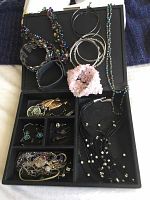 Photo of assortment of costume jewelry including bracelets, earrings and necklaces arranged in a display case with compartments, showing various multi-colored bead necklaces, bracelets in black, silver and pink, along with several pairs of earrings.
