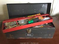 Open black Homak metal toolbox showing assorted tools including hand saw, screwdriver, and packaged hardware inside.