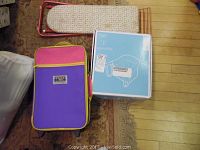 Overview of all items in the lot: small child's iron board, colorful children's suitcase, and boxed Inglesina baby hook on chair.