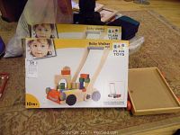 Two boxed Plan Toys wooden baby walkers containing colorful wooden blocks visible on the packaging, placed on carpeted floor next to a wooden lid or tray.