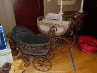 Two vintage baby carriages placed on a wooden floor; the one with black lining and butterfly design is in front, the wicker carriage with a light-colored woven frame behind it.