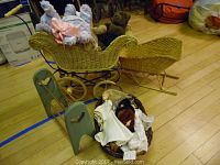 Photo shows large wicker baby carriage with two teddy bears inside, smaller wicker stroller behind, basket with dolls and doll clothes, and green wooden rack in background.