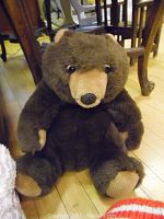 Large brown plush teddy bear sitting on wooden floor near table legs.