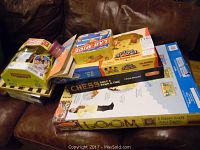 A collection of boxed board games stacked on a couch, showing Monopoly Crazy Cash, Lab Mice, Cuckoo the Rocking Clown, Chess, and Loom craft kit prominently.