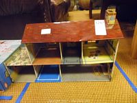 Front view of a small vintage metal doll house with reddish brown shingled roof showing multiple open rooms with different floor colors and wall prints, set on carpeted floor.