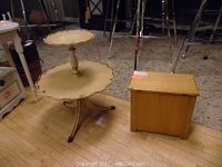Side view of two-tiered yellow toned table with pie crust edges next to rectangular wooden storage box, both on hardwood floor.