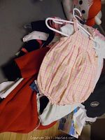 Photo showing a pink and yellow plaid sleeveless romper hanging on a white hanger with multiple other children’s clothing items beneath it, including a red garment and white clothing.