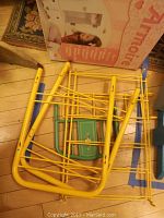 Yellow and blue metal doll bed frame showing spindled side rails, laid flat on wood floor with packaging box in background.