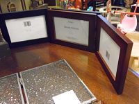 Dark brown trifold wooden picture frame for three photos shown standing on table.