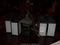 Five outdoor wall lights displayed on a wooden surface including four matching black lantern-style lights with frosted white glass and one vintage metal and colored glass light, showing signs of rust and wear.