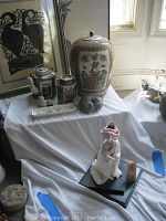 Wide view showing arrangement of china doll, porcelain jars, framed Asian art poster and small decorative items on display with white draped cloth