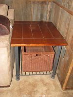 End table viewed at an angle showing wood top, iron legs, and woven basket underneath for storage placed next to a couch and wooden wall.