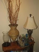 Full view of the decorative items arranged on a wooden surface including decanter, glasses on a tray, basket-style vase with decorative stems, hurricane candle holder, and lamp with brocade shade.