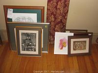 Overall view of six framed artworks leaning against the wall, including a larger black and white 3D print and smaller colorful and monochromatic drawings and paintings.