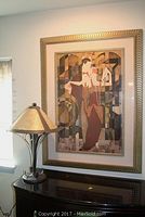 Photo showing an Art Deco style framed print with two figures in period dress alongside a table lamp with metal shade and metal base on a dark surface.