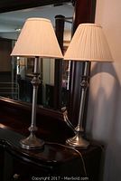 Pair of metal candlestick table lamps shown on a dark wood dresser with mirror, displaying the full lamps including their cream colored pleated shades.