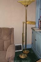 Full view of two floor lamps side by side near couch and cabinet. The taller torchiere lamp has a yellow plastic shade with a brass/gold finish metal base and pole. The shorter adjustable reading lamp is brass/gold with round base and adjustable arm.