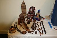 Photo of various African wooden art carvings, including a Noah's Ark boat with paired animals, carved wooden animal figurines, a doll, mask, necklaces, and a woodwind instrument on a white tablecloth.