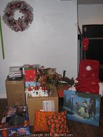 Full view of the lot showing cardboard boxes with snowman figurines, decorative wreath on wall, canvas snowman painting, colorful seasonal pillows, and a chair covered with red Santa hat chair back cover.