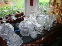 Wide view showing assortment of cut glass and crystal items on a wooden dresser by window.