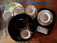 Various porcelain cup and saucer sets with floral designs and gold trim along with a large black bowl and smaller black bowl.
