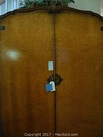 Front view of large wooden armoire with light wood grain finish, ornate metal hardware centered on doors, two doors closed