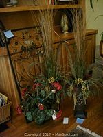 Full view showing matching tall black planters holding faux grasses and flowers, faux geranium plant in planter, and iron metal wall decor leaning against wooden furniture.
