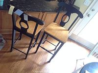 Two wood bar stools with black painted frames and light wood seats shown side by side on a wood floor near granite countertop.