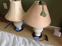 Two white oriental ceramic lamps with different shades, one beige pleated fabric with green tassels and one cream bell-shaped shade, placed on carpeted floor near wall outlet.
