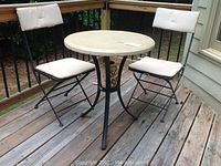 Full set showing round 24 inch diameter cement top table with black metal frame and two black metal folding chairs with white cushions, positioned on a wooden deck.