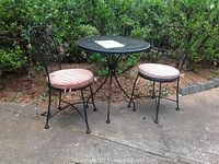 Outdoor black iron metal table with two matching chairs with pink and beige striped round cushions in garden setting.