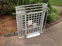 Two white metal child safety gates stacked on pavement outside. Both gates have vertical bars and white metal frames.