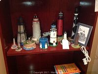 Shelf displaying the collection of 10 lighthouse figurines and lighthouse-themed items, including a candle and framed print, arranged closely together.