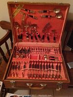 Wide view of open wooden case containing brass flatware including forks, spoons, knives, and serving pieces, arranged on orange felt lining.