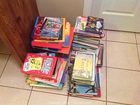 Multiple stacks of children's reading books placed on tile floor near a wooden cabinet and door, showcasing variety of book sizes and covers.