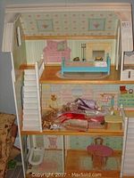 Front view of three-level wooden dollhouse with stairs, various doll furniture pieces including pink armchair, blue bed, and assorted doll clothes visible on middle level.