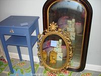 Blue side table next to two mirrors, one with ornate gold plastic frame, one rectangular with wooden frame